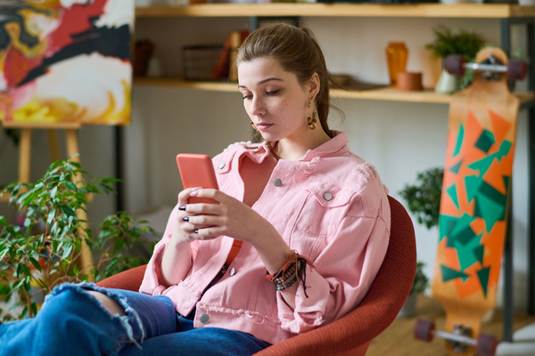 A young woman with gathered hair in a pink denim jacket with bracelets on her arm is sitting in a red armchair and using her phone